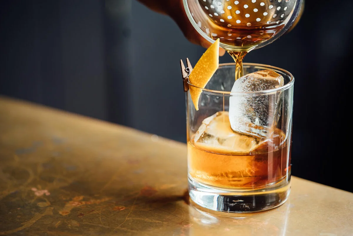 Bartender pouring an Old Fashioned cocktail over ice with an orange peel garnish.
