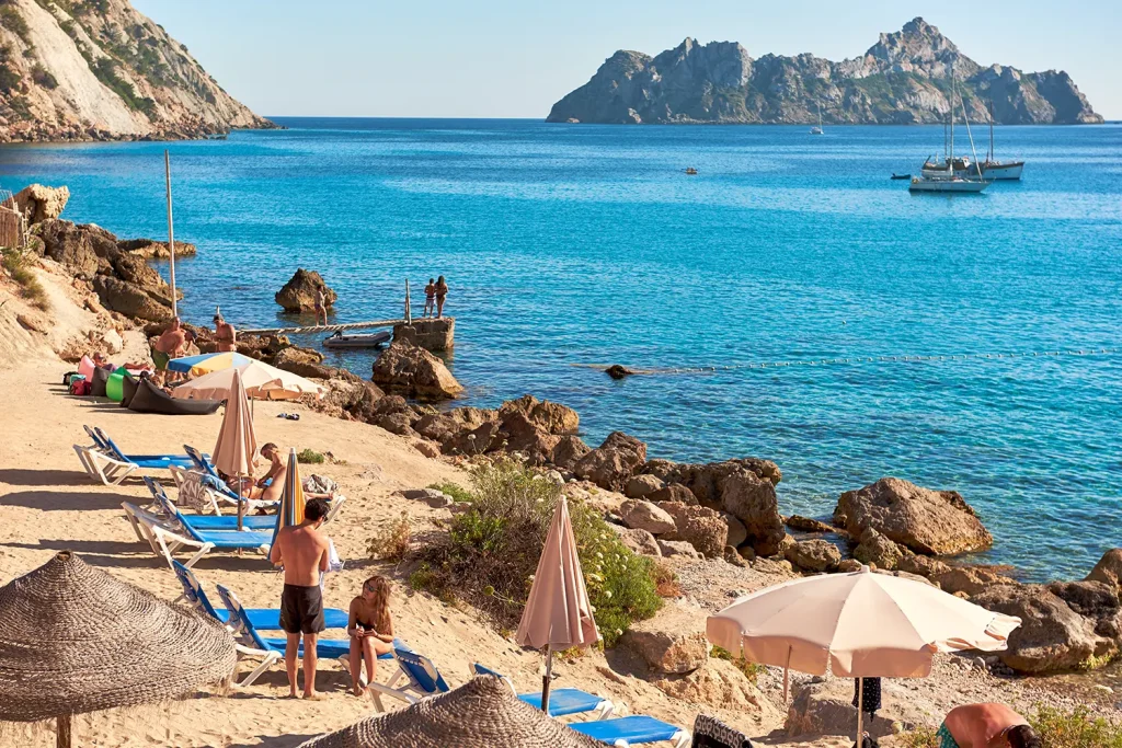 Ibiza beach with turquoise waters, rocky cliffs, and sun loungers by the shore.