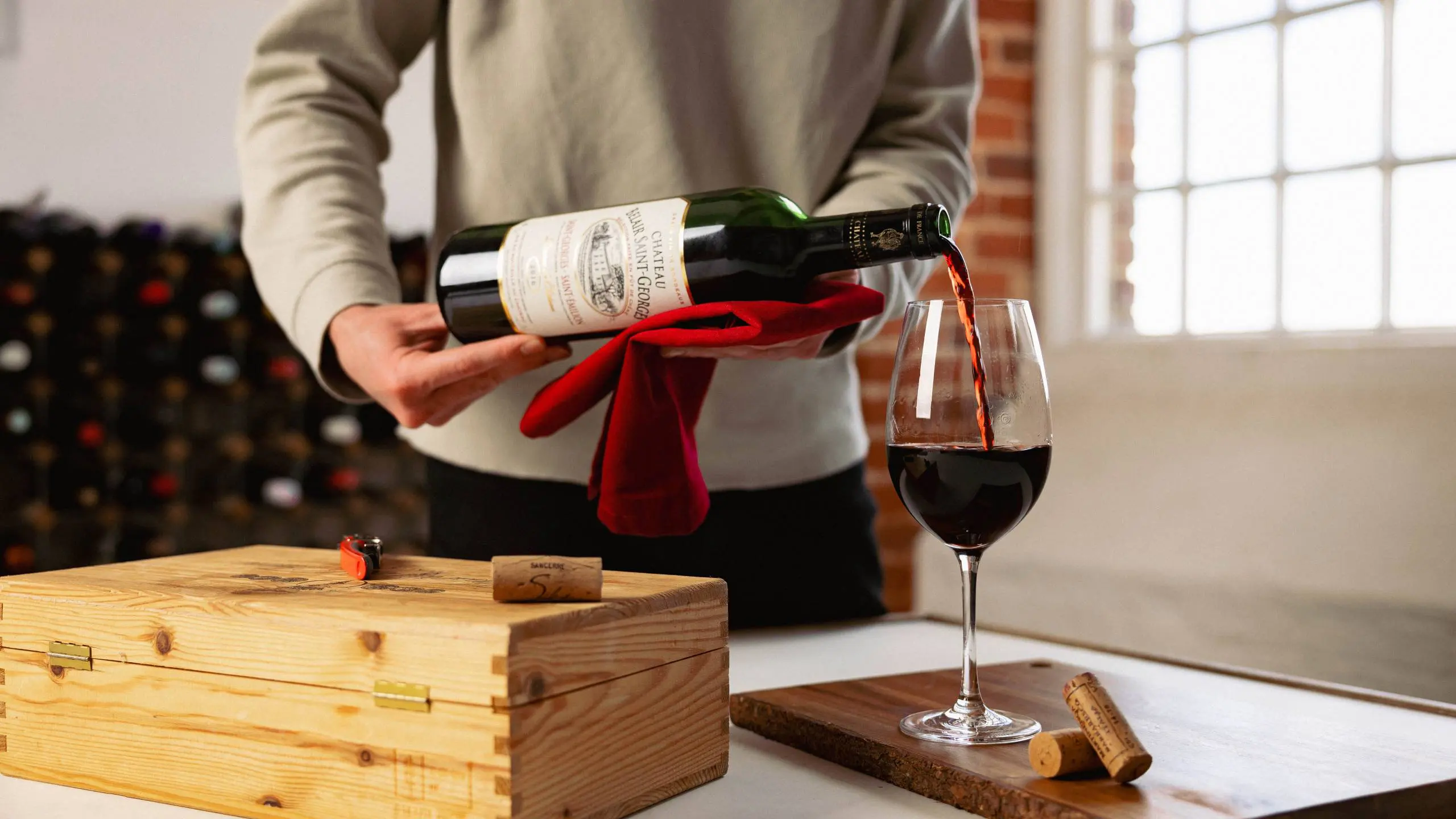 Person pouring red wine from a bottle into a wine glass beside a wooden box.