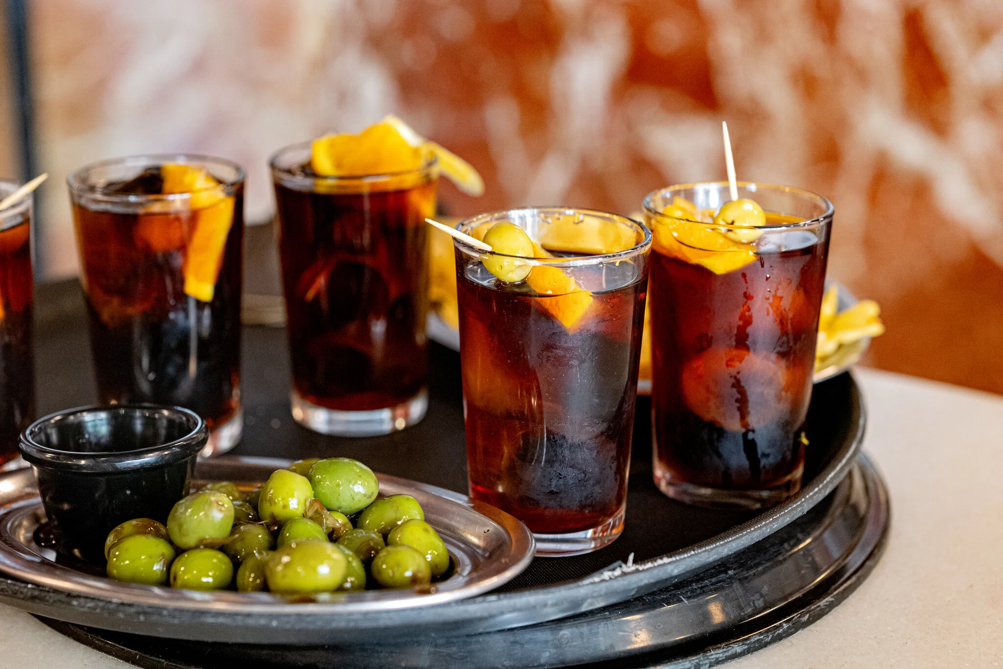 Glasses of dark vermouth with orange garnishes and olives on a tray.