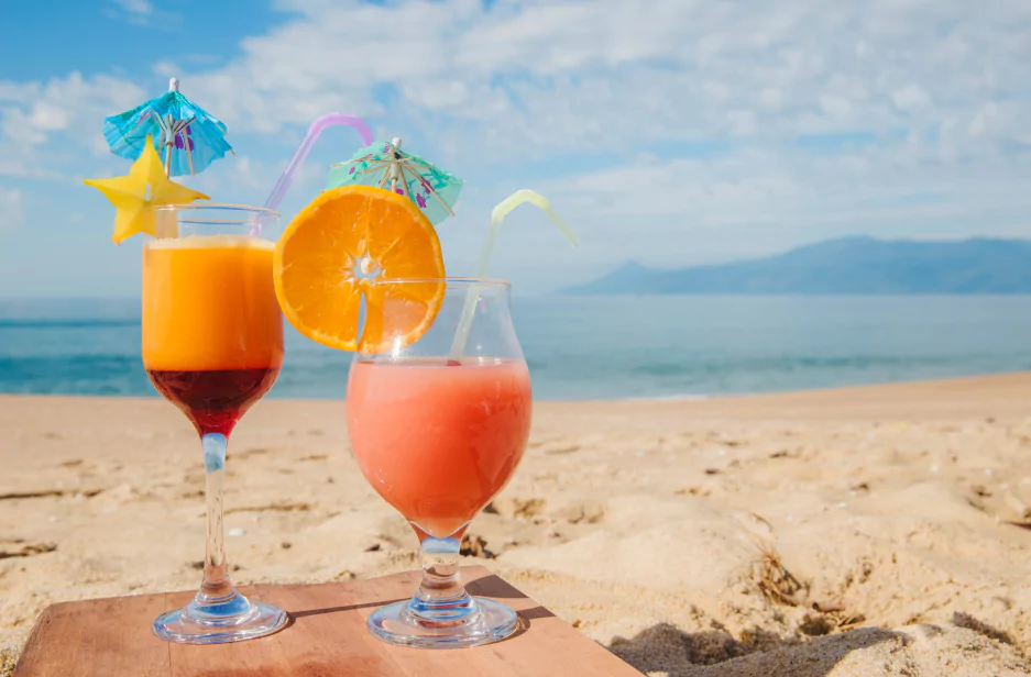 Two colorful tropical drinks on a sunny beach with the sea in the background.