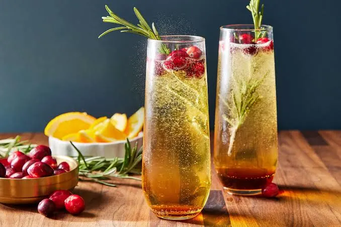 Two sparkling holiday drinks with rosemary, cranberries, and citrus garnish on a wooden table.