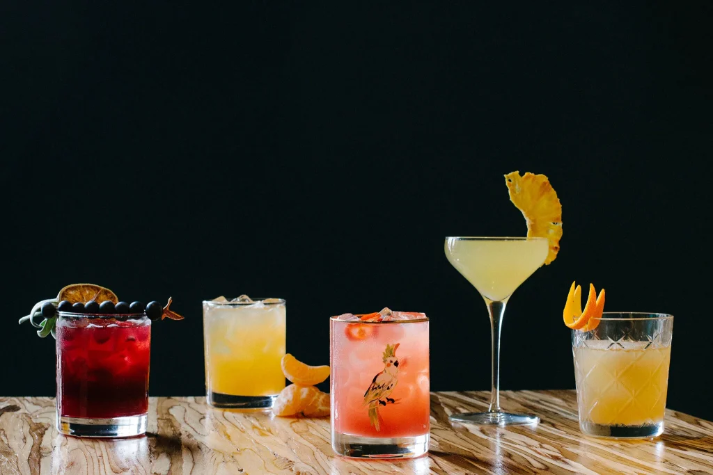 Assorted colorful cocktails with fruit garnishes on a wooden bar.
